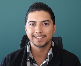 Hombre joven de tez morena sonriendo frente a un fondo verde oscuro.